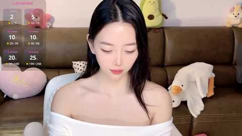 Media: Video of an East Asian woman with fair skin, long black hair, and a slender physique, wearing a white off-shoulder top, sitting on a brown leather couch. Background includes stuffed toys and a digital screen displaying weather information.