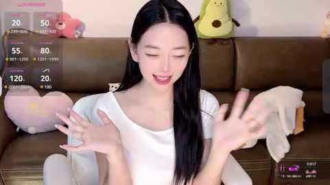 Media: Video of a smiling Asian woman with long black hair, wearing a white T-shirt, sitting on a brown couch, surrounded by plush toys.