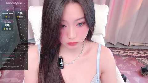 Media: Video of a young Asian woman with long, straight black hair, wearing a light blue bra and silver necklace. She sits on a bed with pink and brown bedding, in a room with soft lighting.