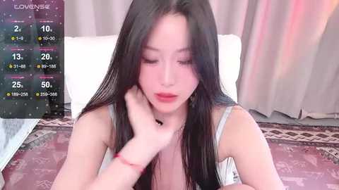 Media: Video of an Asian woman with long black hair, wearing a light grey tank top, sitting on a patterned rug, with a smartphone in front of her displaying a live stream overlay.