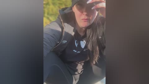 Media: A video captures a young woman with fair skin, wearing a black jacket, a black beanie, and sunglasses, with a blurred green background. She is sitting outdoors, shielding her eyes from the sun.