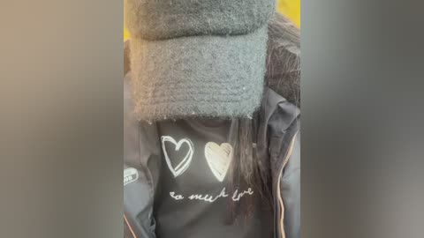 Media: Video of a person wearing a grey beanie, black jacket with white heart and \"love\" text, and a grey scarf, partially obscured by the beanie, against a blurred yellow background.