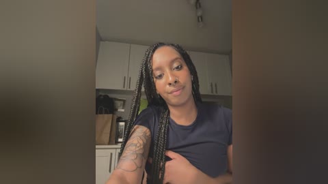 Media: Video of a young African-American woman with long braids, wearing a dark t-shirt, standing in a kitchen with white cabinets, black cat, and a brown bag.