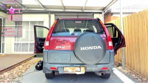 Media: Video of a red Honda SUV with a black spare tire cover, parked in a driveway with a wooden fence. The SUV's rear door is open, revealing the interior. The background features a beige house with a large window and a sign in the upper left corner.