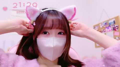 Media: Video of a young Asian woman in a pink plush cat headband, white face mask, and furry pink robe, adjusting her cat ears, in a brightly lit room with pink decorations and a calendar.