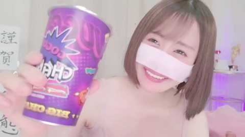 Media: Video of an Asian woman with straight brown hair, holding a purple \"Chupa Chups\" lollipop, wearing a white tape over her mouth, smiling, in a bedroom with pastel decor and a pink bedspread.