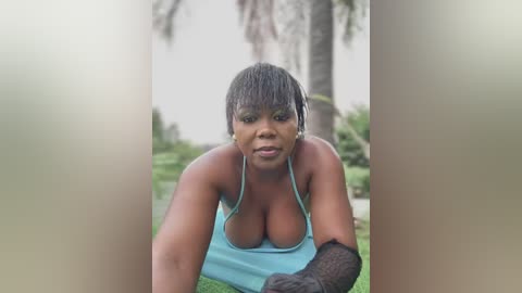 Media: Video of an African-American woman with medium-dark skin, short black hair, and a light blue sports bra, crouching in a lush garden with palm trees and greenery in the background.