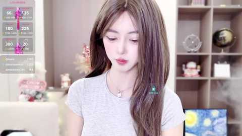 Media: Video of a young Asian woman with long brown hair, wearing a grey t-shirt, indoors with a shelf filled with decorative items and a TV screen in the background.