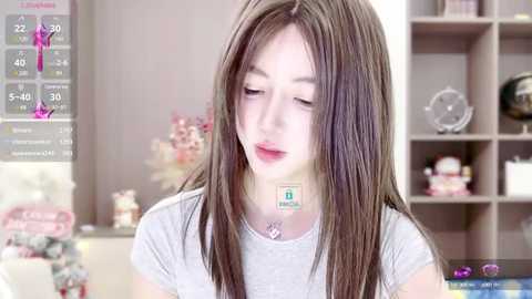 Media: Video of a young woman with straight, light brown hair, wearing a gray t-shirt, in a softly lit, modern bedroom with white shelves and decorative items.