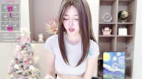 Media: A video of an Asian woman with long, straight brown hair, wearing a white crop top, standing in a modern living room with a Christmas tree, bookshelf, and a Starry Night painting.