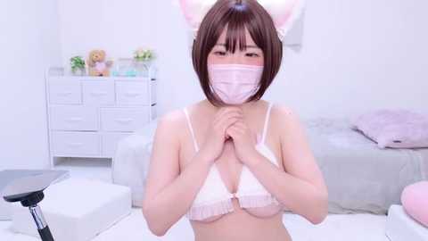 Media: Video of an Asian woman with short brown hair, wearing a pink face mask, bunny ears, and a white lace bra, standing in a white bedroom with a bed, dresser, and plush toys.