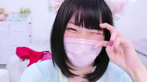 Media: Video of an Asian woman with short black hair, wearing a pink mask, cat ears, and light blue top, winking, in a bright, white room with pink and white decor.