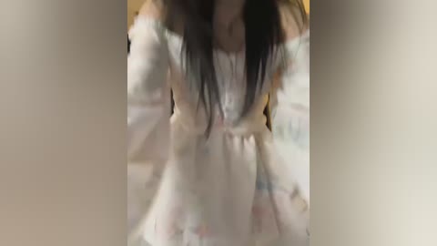 Media: Video of a woman in a white, off-the-shoulder dress with long, dark hair, standing indoors with a blurred background.