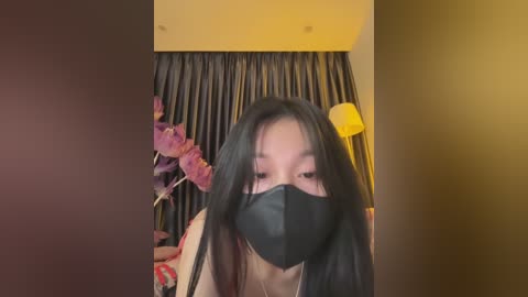 Media: Video of an Asian woman with long black hair, wearing a black mask, in a dimly lit room with floral decor and yellow lighting.