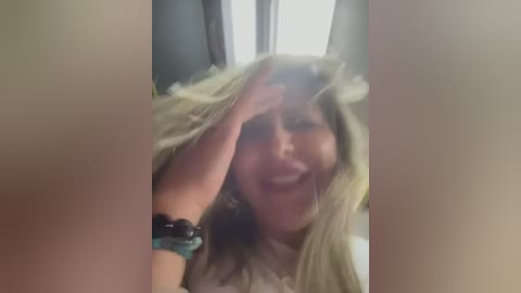 Media: A blurry video shows a woman with long blonde hair, wearing a white top and a black beaded bracelet, smiling and covering her face with her hand.