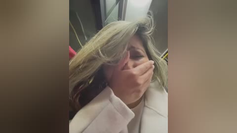 Media: Video of a woman with gray hair, crying, hiding her face with her hand, wearing a white coat, in a dimly lit, possibly public, setting.