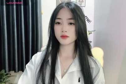 Media: Video of an East Asian woman with long black hair, wearing a white shirt, standing indoors. She has fair skin, a slender physique, and red lipstick. Background features a dark curtain, white wall, and a plant.