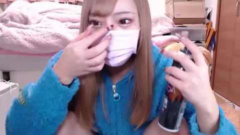 Media: Video of an Asian woman with long, light brown hair and fair skin, wearing a blue fuzzy sweater, face mask, and holding a can of hairspray and a comb. She's in a messy bedroom with a bed and boxes in the background.