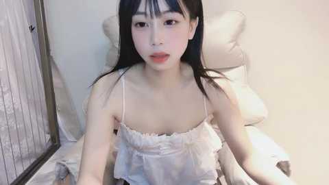 Media: Video of an East Asian woman with straight black hair and pale skin, wearing a white, frilly camisole. She sits on a bed with white pillows, indoors.