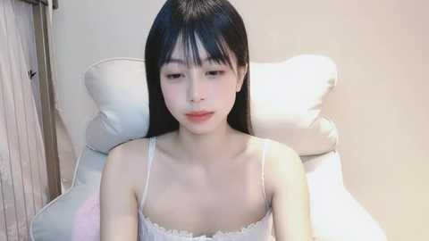 Media: Video of a young Asian woman with straight black hair, fair skin, and delicate facial features, wearing a white lace-trimmed spaghetti strap top, seated on a white chair in a well-lit room with beige walls.