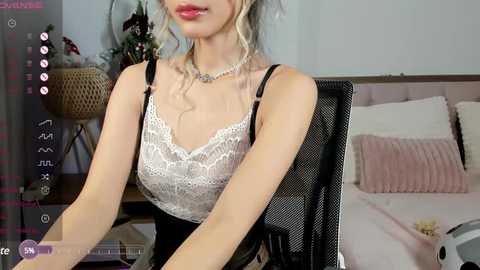 Media: Video of a fair-skinned woman with wavy blonde hair, wearing a white lace bra and black suspenders, sitting on a black chair in a cozy bedroom.