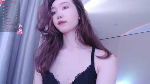 Media: Video of a fair-skinned Asian woman with long, wavy brown hair, wearing a black lace bra, standing indoors with a grey curtain and a partially visible wall clock in the background.