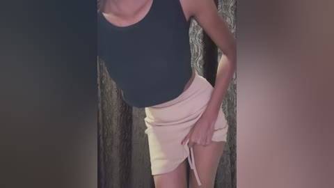 Media: Video of a slim, light-skinned woman with a small bust, wearing a dark green tank top and light beige shorts, adjusting her shorts' waistband. She stands against a textured dark curtain backdrop.