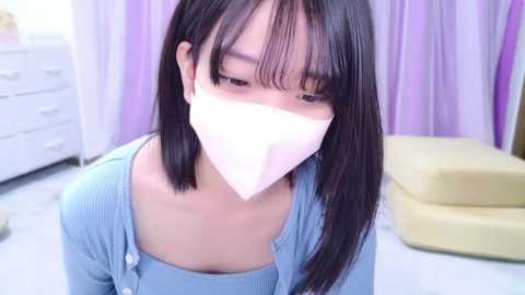 Media: Video of an Asian woman with straight black hair, wearing a light blue top, a white surgical mask, and light purple curtains in the background.