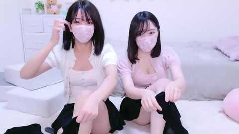 Media: Video of two Asian women with short black hair, wearing masks, white tops, and black skirts, sitting on a white bed, adjusting their masks, in a bright, minimalist bedroom.