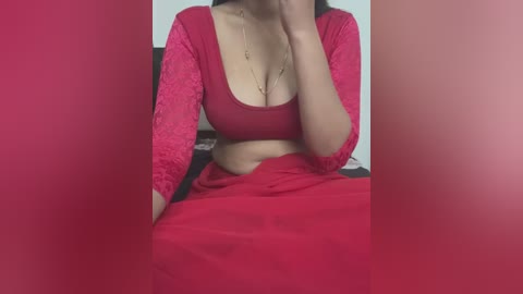 Media: Video of a woman in a red lace top, revealing cleavage, and red skirt, sitting on a bed, with a blurred background, hand covering her mouth.