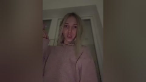 Media: Video of a blonde woman with fair skin, wearing a light pink sweater, standing in a dimly lit room with white walls and a window.