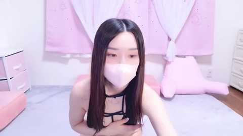 Media: A video of an Asian woman with long black hair and light skin, wearing a black bra, pink mask, and pink panties, kneeling on a bed with pink and white bedding.