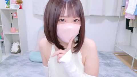 Media: Video of a young East Asian woman with straight brown hair, wearing a white sleeveless top, light pink mask, and finger to lips, in a soft-lit, minimalist bedroom with white furniture and pastel decor.