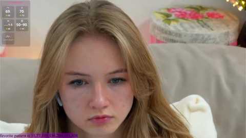 Media: Video of a young Caucasian woman with long blonde hair and blue eyes, wearing a white sweater, sitting on a beige couch in a well-lit room with a floral-patterned cushion in the background.