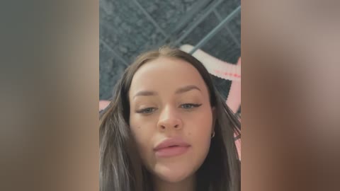 Media: Video of a young woman with fair skin, straight brown hair, and light makeup, standing in a bathroom with black and white tiled walls and a pink towel.