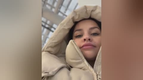 Media: A video of a young woman with light skin and dark hair, wearing a beige puffer jacket, slightly covering her face, against a blurry industrial background.