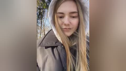 Media: Video of a young woman with long, straight blonde hair, wearing a grey hoodie, looking downcast in a forest setting with tall trees and a blue sky.