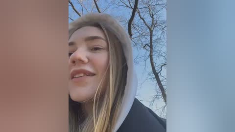 Media: Video of a young Caucasian woman with straight blonde hair, light skin, and a slight smile, taken outdoors against a backdrop of bare trees and a clear blue sky.