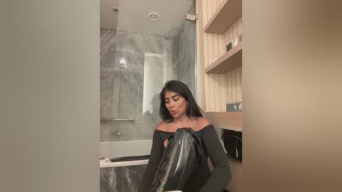 Media: Video of a woman with medium brown skin, wearing a black off-shoulder top, sitting in a modern bathroom with a glass shower, beige tiles, and wooden shelves.