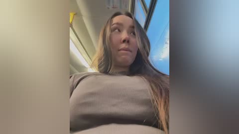 Media: Video of a woman with long brown hair, wearing a light brown sweater, looking out a train window at a clear blue sky.