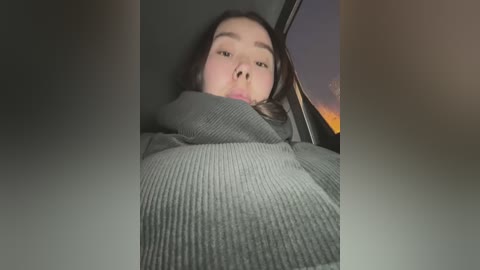 Media: A video of a young Asian woman with long black hair, wearing a gray ribbed sweater, lying in the backseat of a car. The window shows a sunset with orange and purple hues.