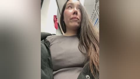 Media: Video of a young woman with long brown hair, wearing a gray shirt and green jacket, looking contemplative in a subway car with red seats and white walls.