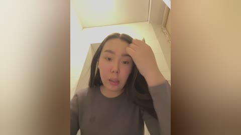 Media: Video of a young woman with fair skin, straight brown hair, and light makeup, wearing a gray top, standing in a narrow hallway with beige walls, looking slightly confused with hand on head.