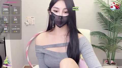 Media: Video of an East Asian woman with long black hair and fair skin, wearing a gray off-shoulder top, black face mask, and dangling earrings, sitting at a desk with a plant, digital clock, and white wall background.