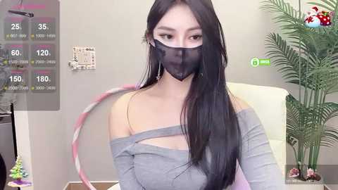 Media: Video of an East Asian woman with long black hair, wearing a gray off-shoulder top, black face mask, and pink hoop earrings, seated in a modern, minimalistic room with a green plant and white furniture.