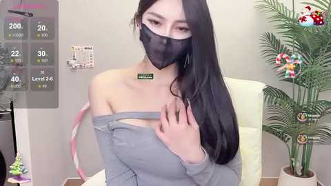 Media: Video of an Asian woman with long black hair, wearing a gray off-shoulder sweater, black mask, and a green necklace. She's indoors, in front of a white wall, with a potted plant and a digital display showing health metrics in the background.