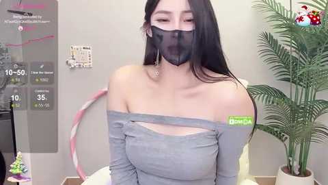 Media: A video of an East Asian woman with long black hair, wearing a gray off-shoulder top, black face mask, and pink hoop earrings, standing in a room with a potted plant and white wall.