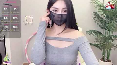 Media: Video of an Asian woman with long black hair, wearing a gray off-shoulder top, black mask, and holding a phone, standing in a room with a green plant and a digital display.