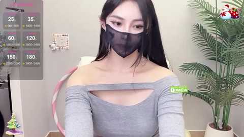 Media: Video of an Asian woman with long black hair, wearing a grey off-shoulder top, black mask, and green name tag, standing in a room with a potted plant and white wall.