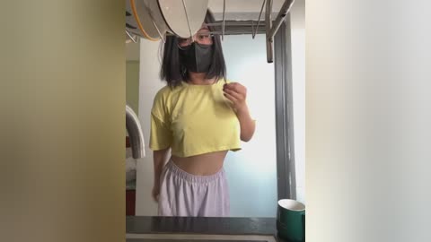 Media: Video of a young Asian woman with straight black hair, wearing a yellow crop top, black mask, and loose pink shorts, taking a selfie in a narrow, white-walled hallway.